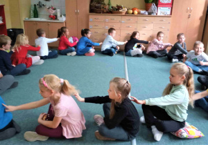 Uczniowie siedzą w kręgu na dywanie w sali lekcyjnej. Zwróceni są do siebie plecami i wykonują relaksujące masażyki.
